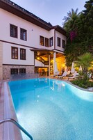 A pool area with lounge chairs and umbrellas, a stone building with multiple windows, and a well-lit interior.