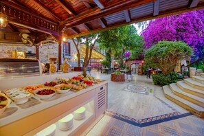 Ein Bereich mit Buffet und verschiedenen Speisen, sowie Sitzgelegenheiten mit Blick auf lila Blumen und eine Fontäne.