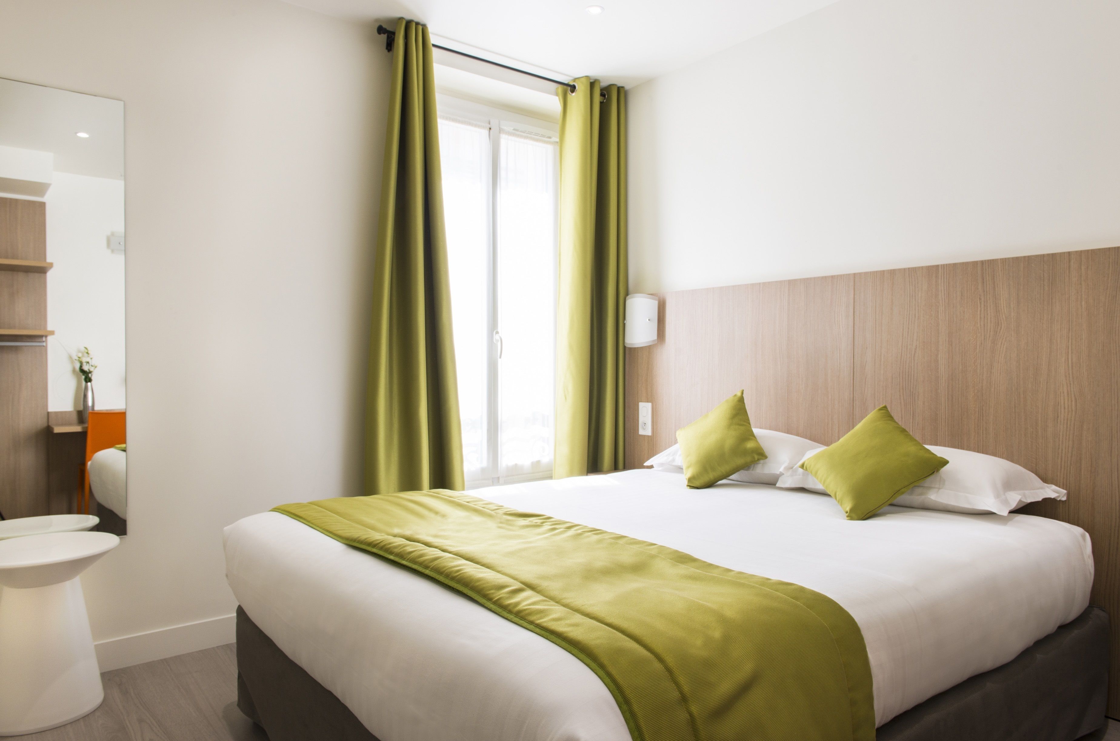 A hotel room with a large bed, green curtains, a wooden headboard, a white bedside table, and a mirror.