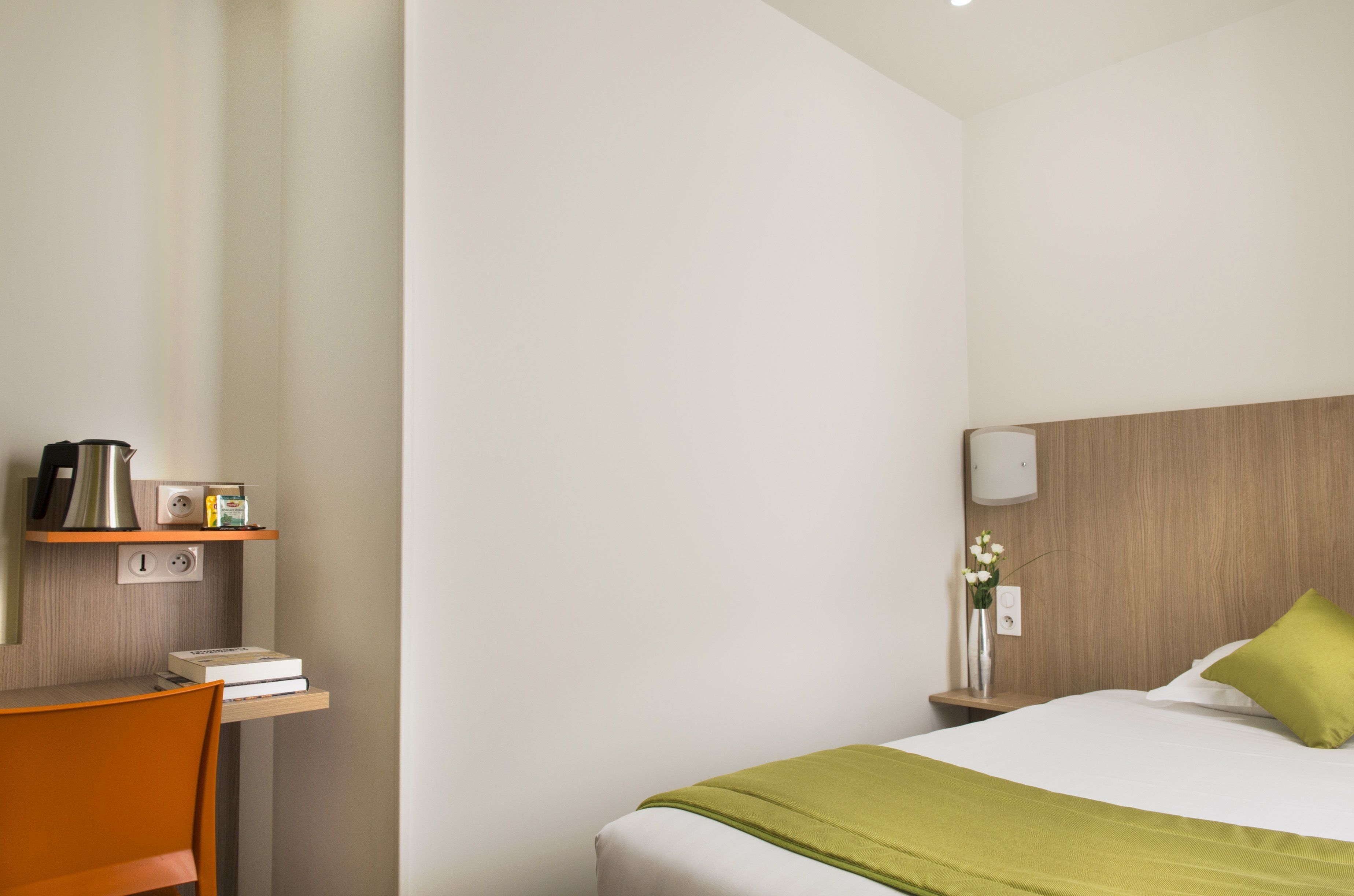 A hotel room with a bed, a desk with a chair, a nightstand with a lamp, and a wall-mounted shelf with a kettle and books.