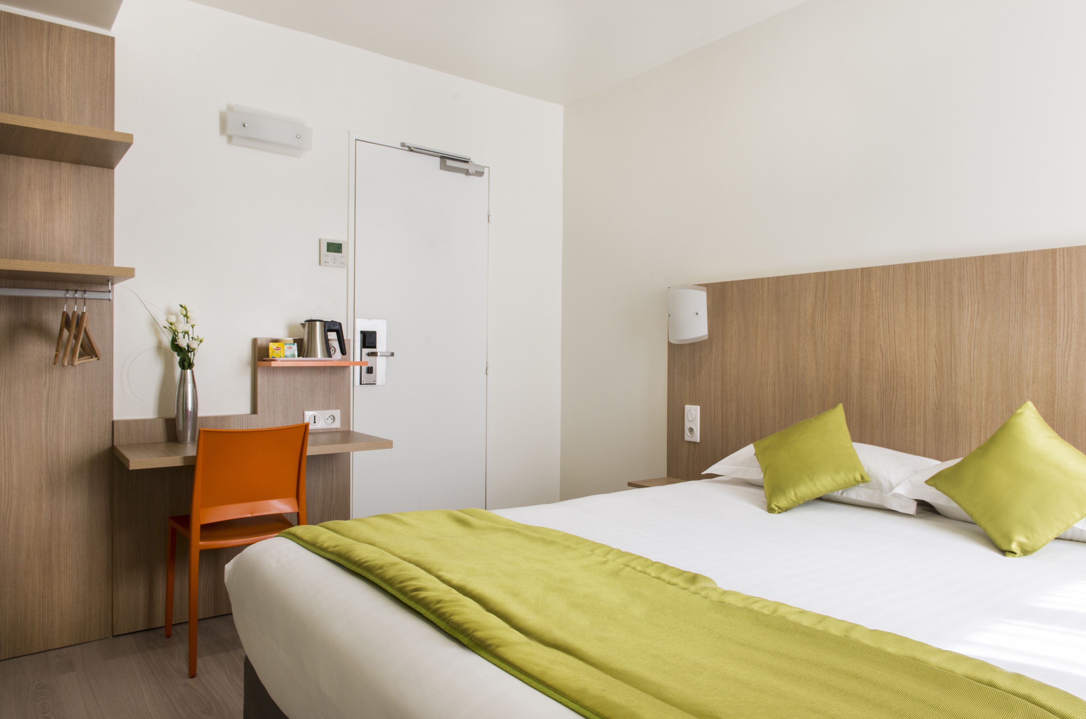 A hotel room with a bed, a desk, an orange chair, a green throw blanket, and a wooden headboard.