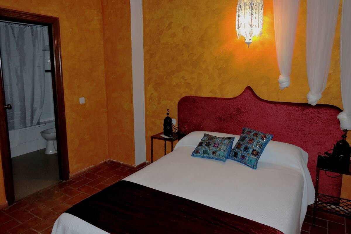 A bedroom with a red headboard, a white bedspread, two patterned pillows, a nightstand with a lamp, and a wall-mounted light fixture.