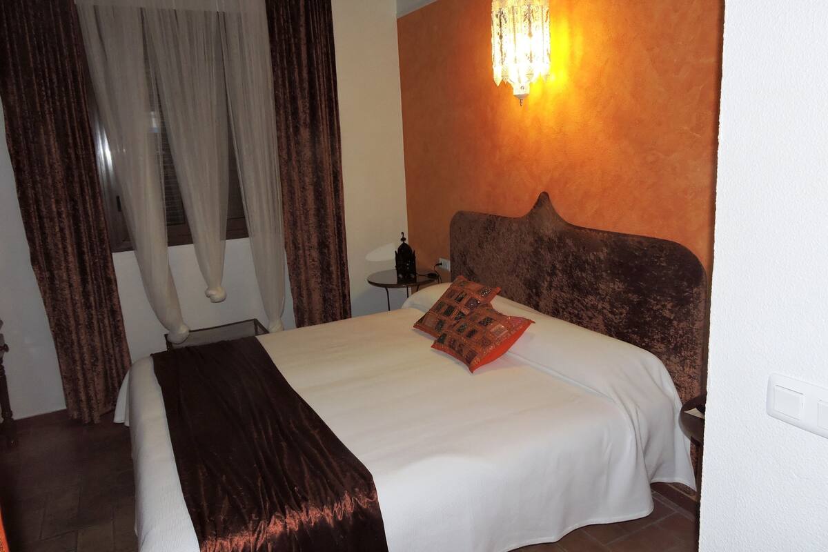 A hotel room with a large bed, a nightstand, a wall-mounted lamp, and a window with curtains.