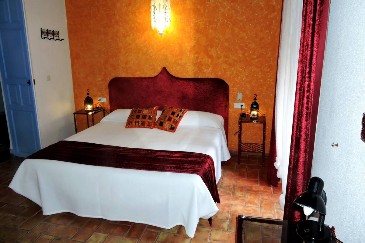 A hotel room with a large bed, two bedside tables with lamps, an orange textured wall, and a blue door.