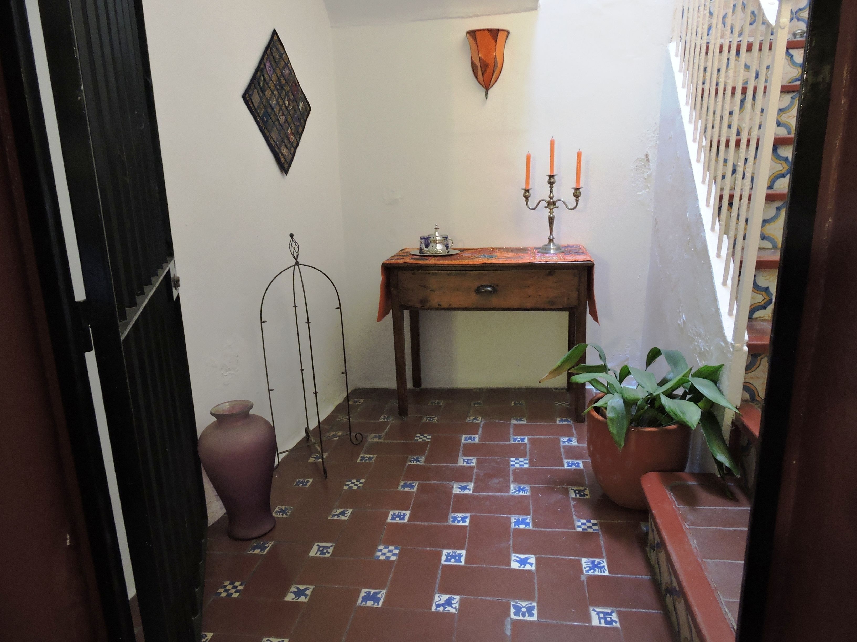 Un pasillo estrecho con suelo de baldosas, una pequeña mesa con un candelabro, una planta en maceta y un adorno en la pared.