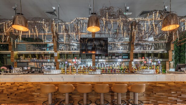 En bar med et ternet gulv, flere barstole samt diverse flasker og glas på hylderne.