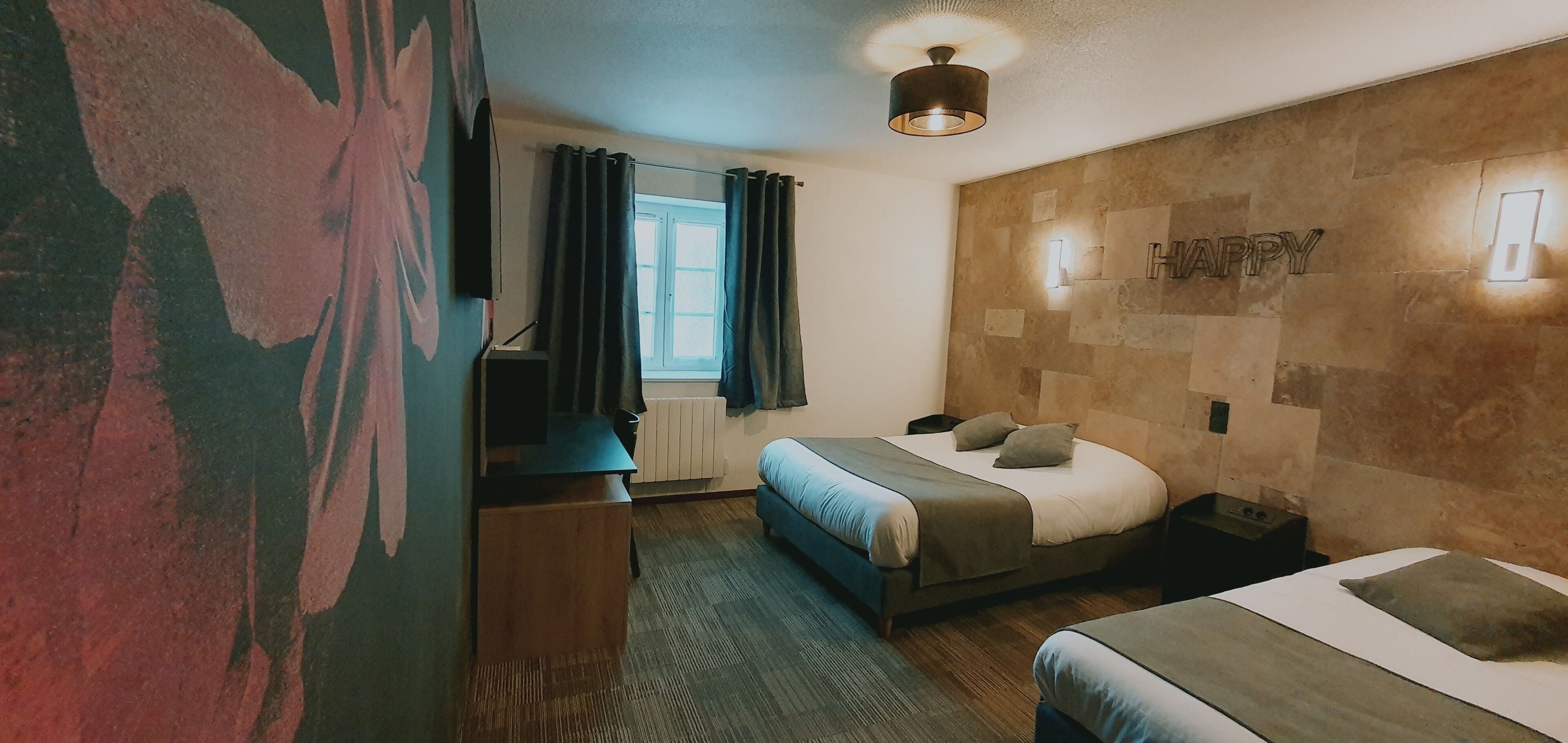 A hotel room with a bed, a desk, a chair, a window with curtains, a wall-mounted light, and a ceiling fan.