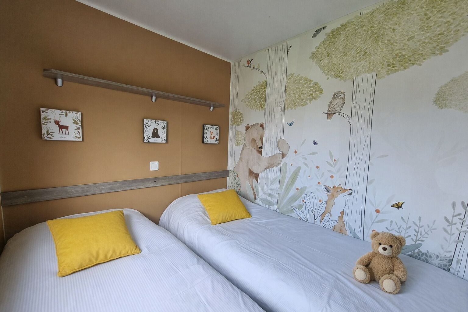 Une chambre avec deux lits, chacun orné d’un oreiller jaune, et un ours en peluche placé sur le côté droit.