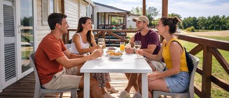 A group of people dining at a wooden deck table with a view of a house and greenery in the background.