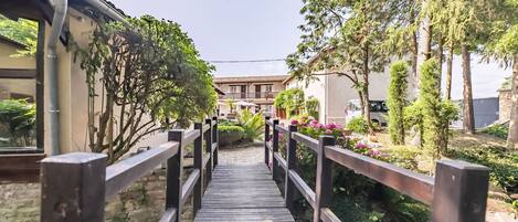 A wooden bridge leading to a building with a tiled roof, surrounded by greenery and a parked car.
