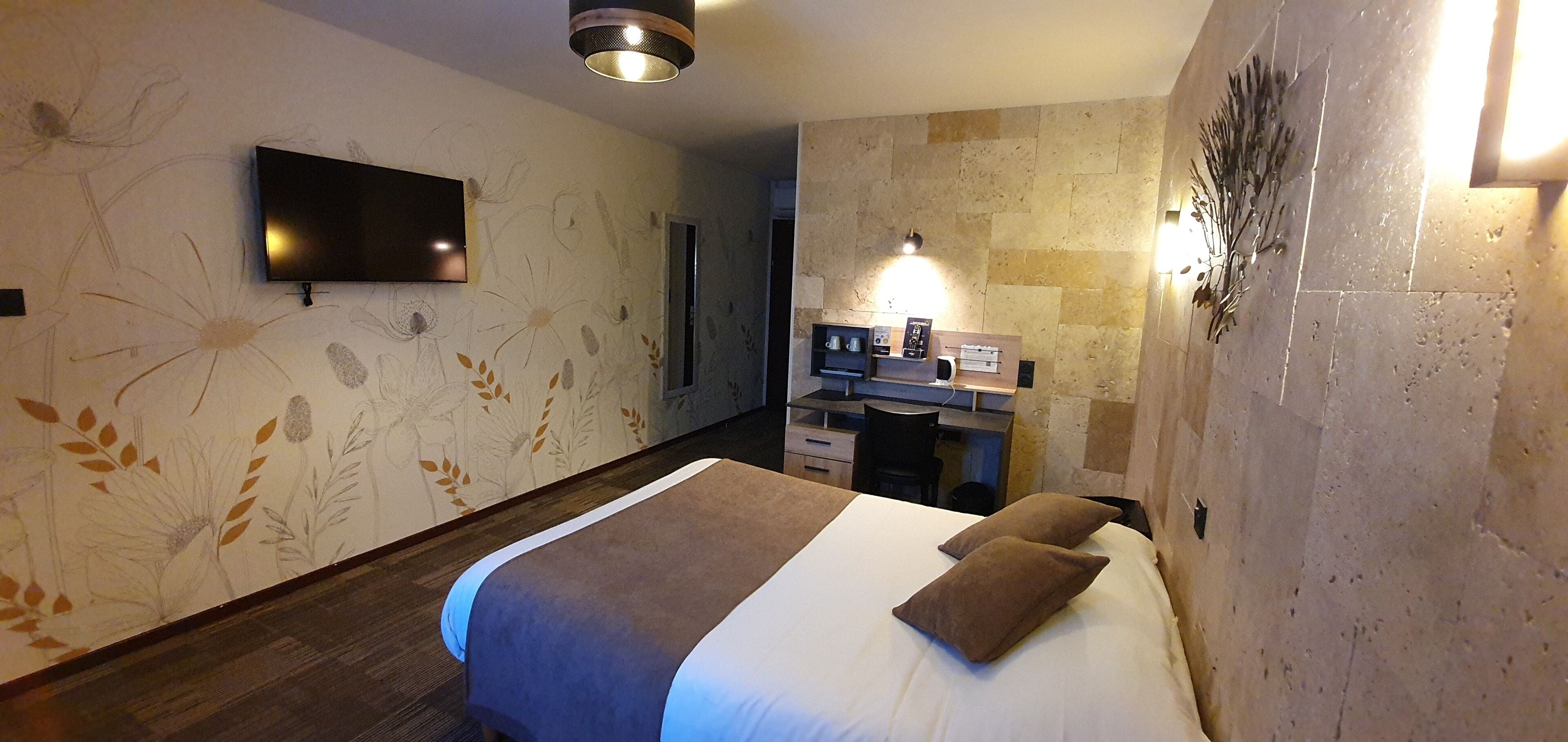 A hotel room with a bed, a flat-screen TV mounted on the wall, a desk with a chair, and a decorative wall piece.