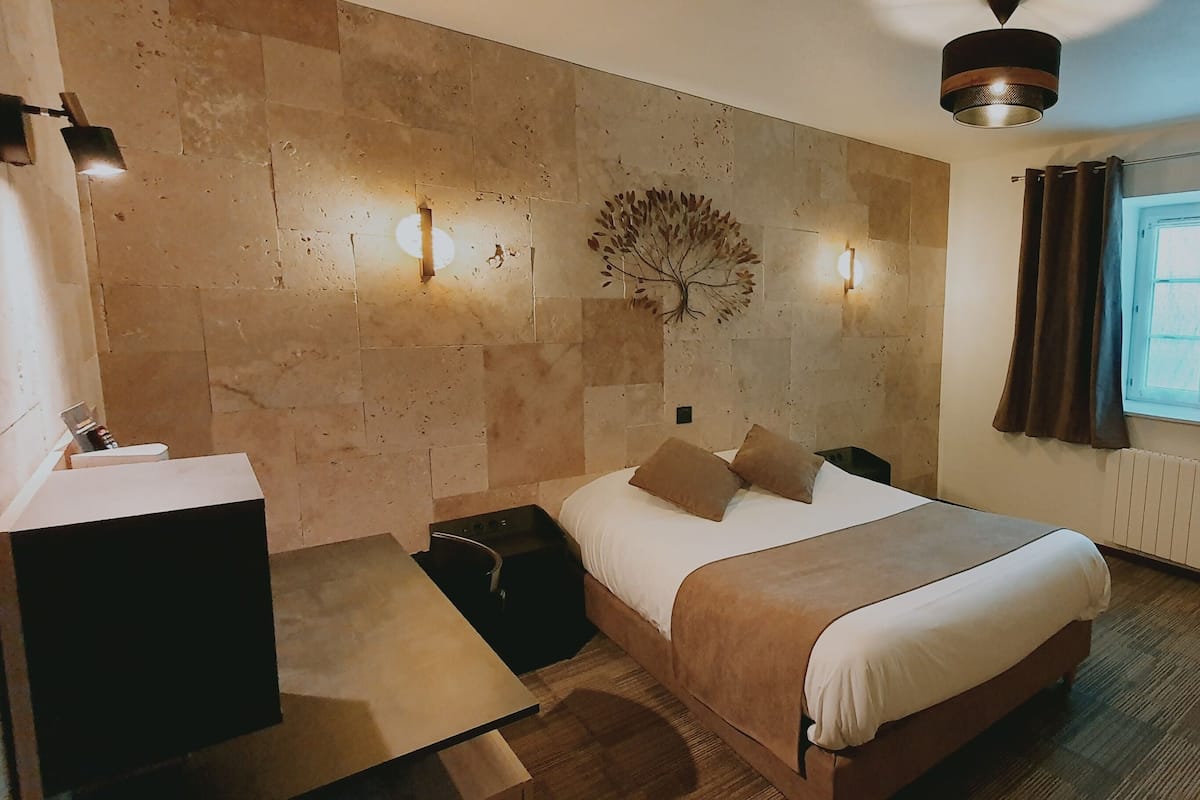 A hotel room with a bed, bedside table, lamp, and a wall-mounted tree decoration.