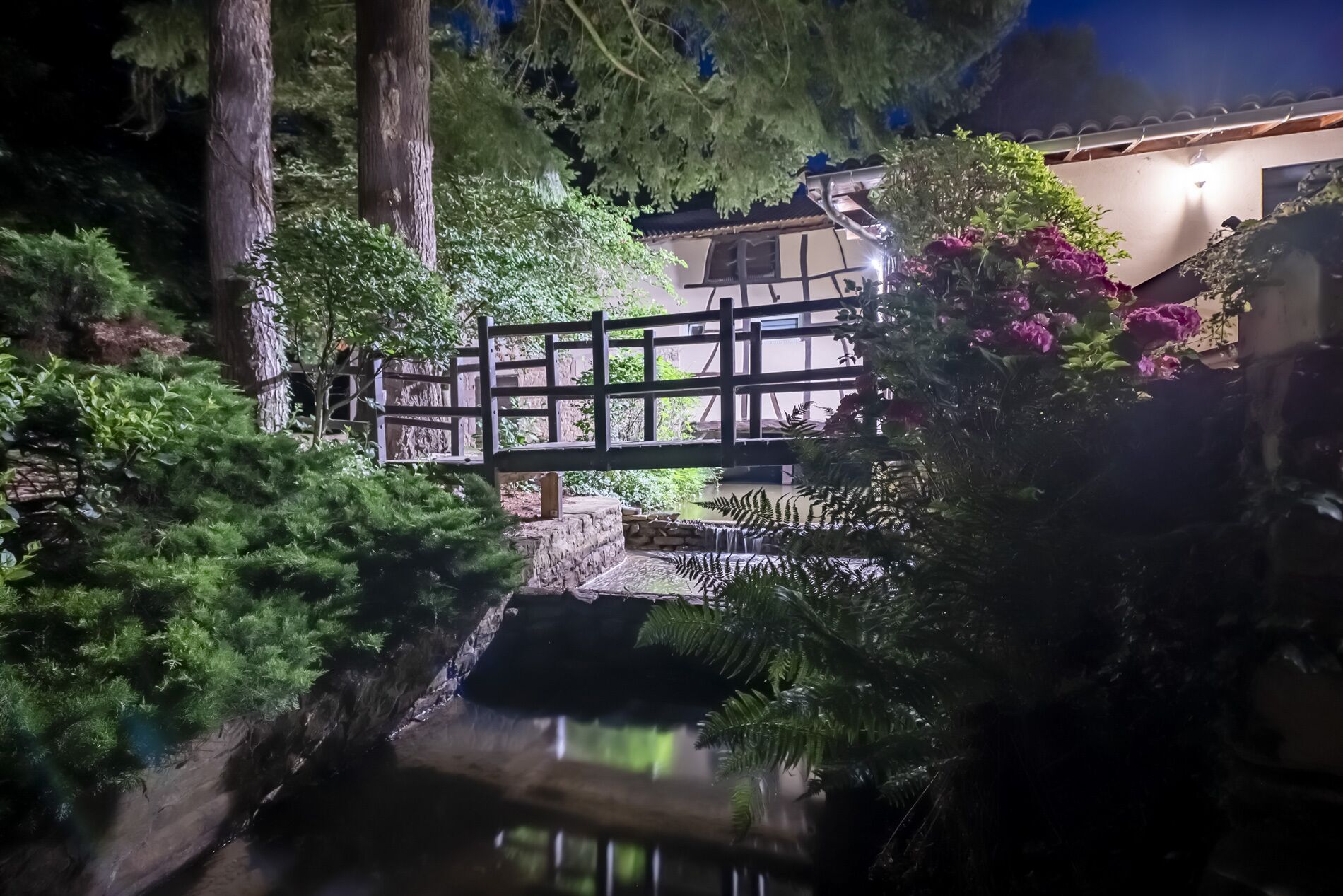A traditional Japanese-style garden with a wooden bridge over a small stream, surrounded by lush greenery and flowering plants.