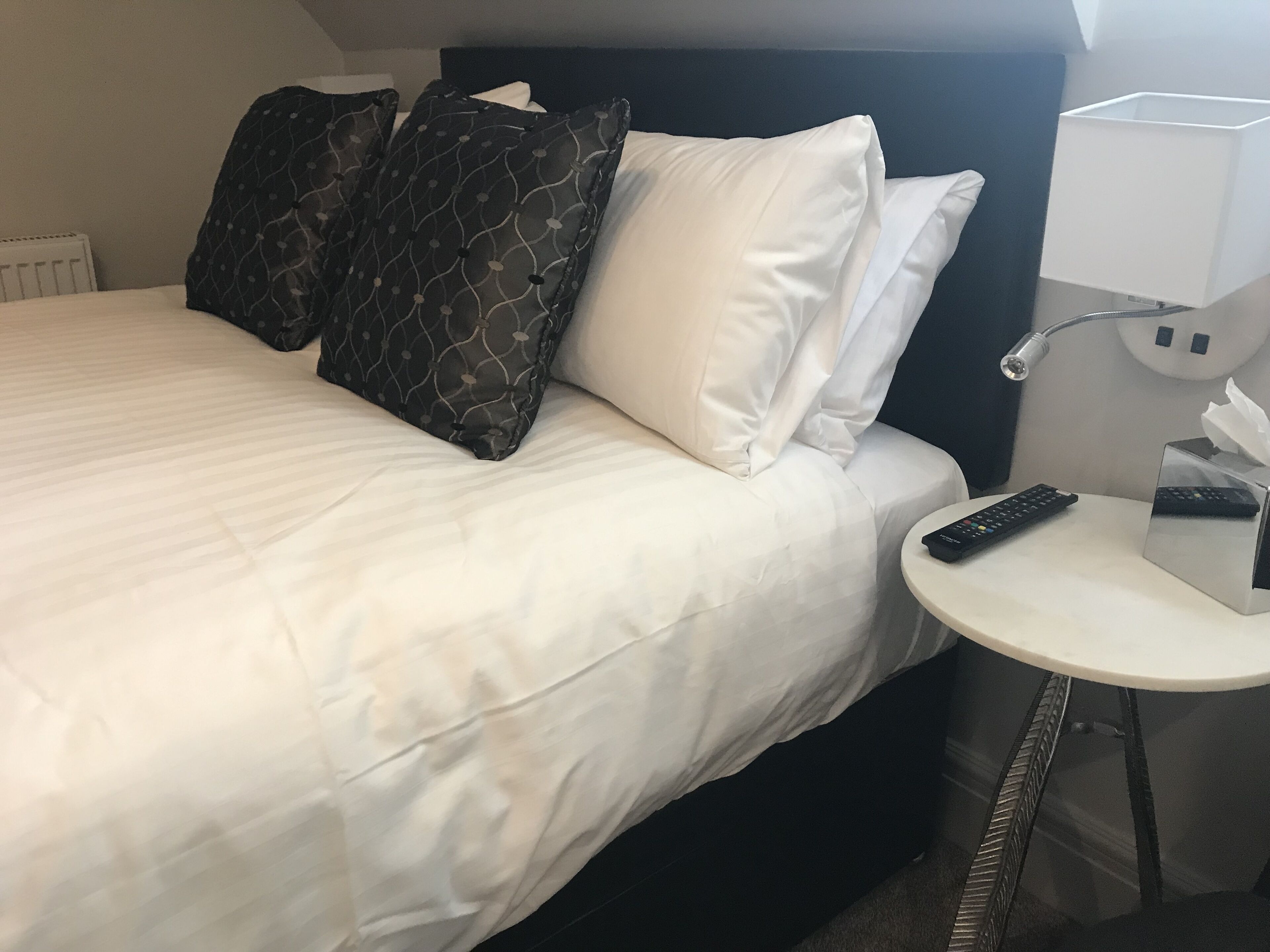 Habitación de hotel con cama, almohadas, mesita de noche con control remoto y lámpara.
