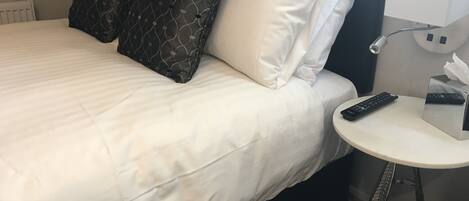 Habitación de hotel con cama, almohadas, mesita de noche con control remoto y lámpara.