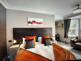 A modern bedroom with a large bed, a cityscape artwork, a desk with a lamp, and a view of greenery through the window.