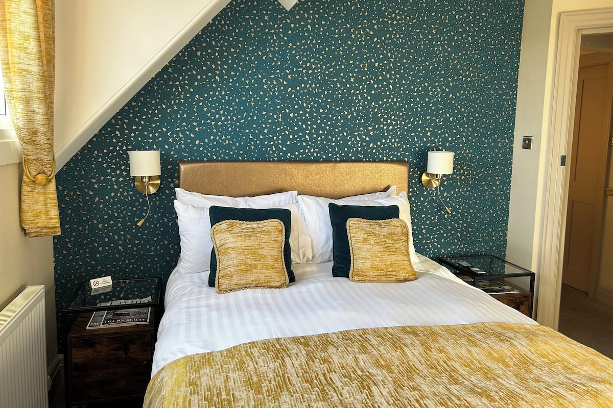 Un dormitorio con techo inclinado, cama con ropa de cama blanca y dorada, dos lámparas fijadas a la pared y una pared verde oscuro con estampado.
