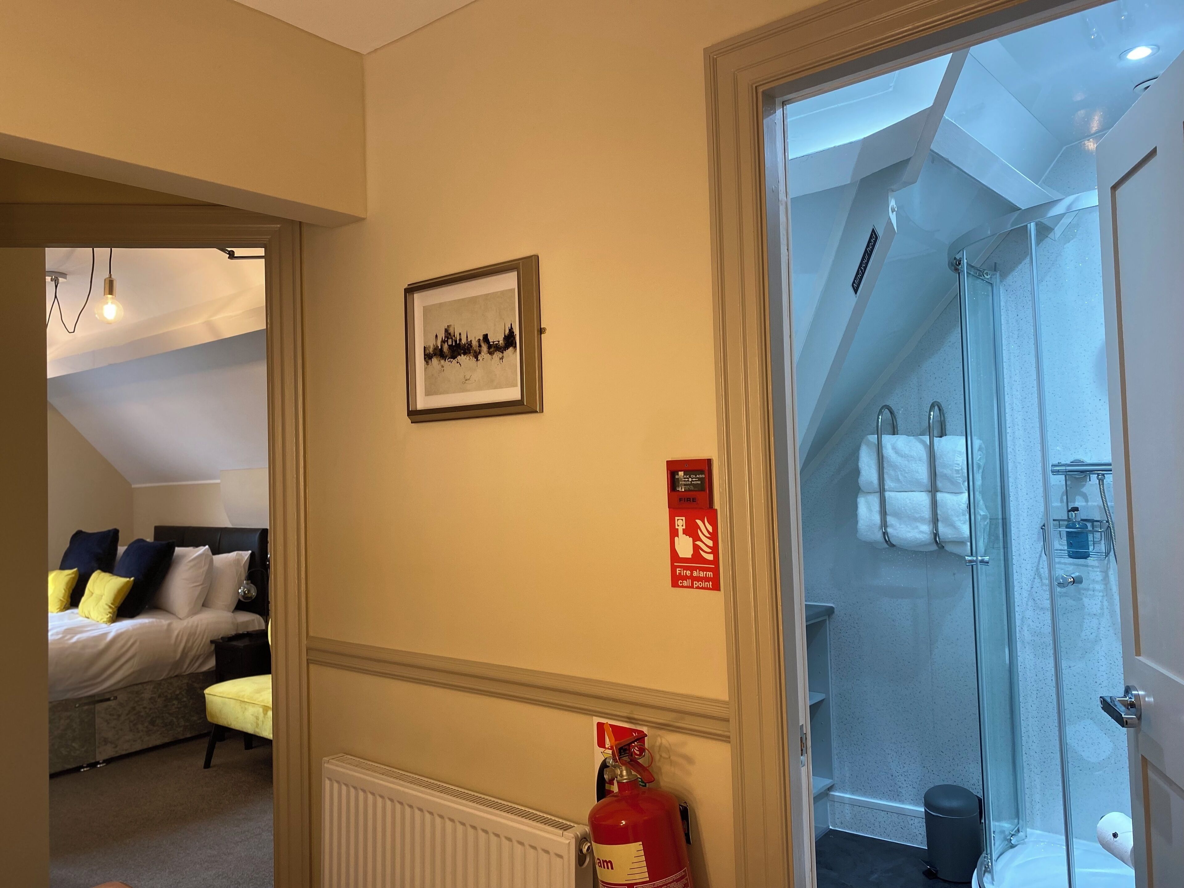 A hotel room with a bed, a fire alarm call point, and a shower area.
