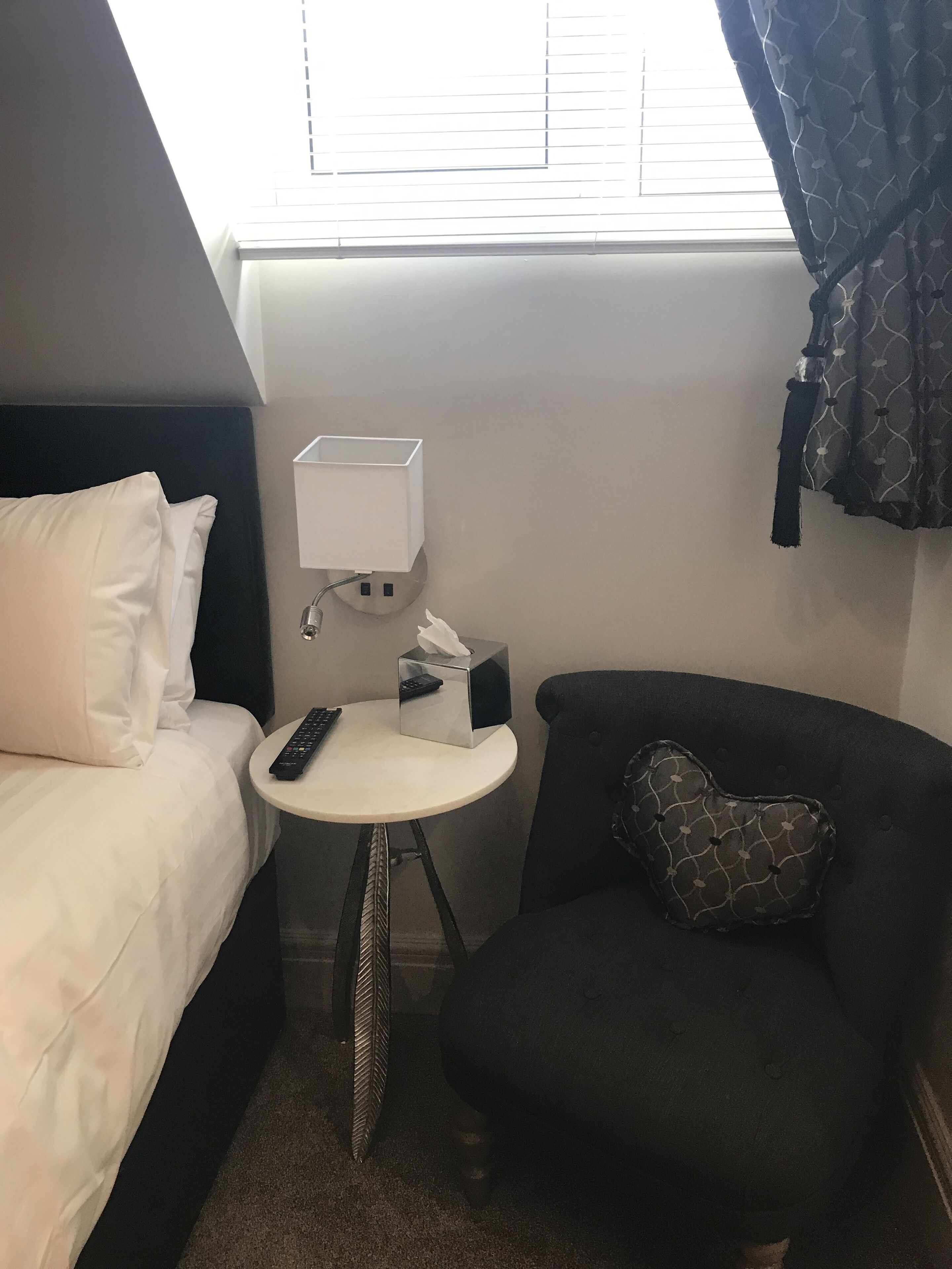 Une chambre d’hôtel avec un lit, une petite table ronde sur laquelle se trouve une télécommande, une boîte de mouchoirs et une chaise avec un coussin à motifs.