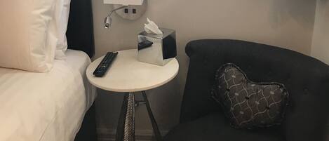 Hotelkamer met een bed, een klein rond tafeltje met een afstandsbediening, een zakdoekendoos en een stoel met een gestreept kussen.