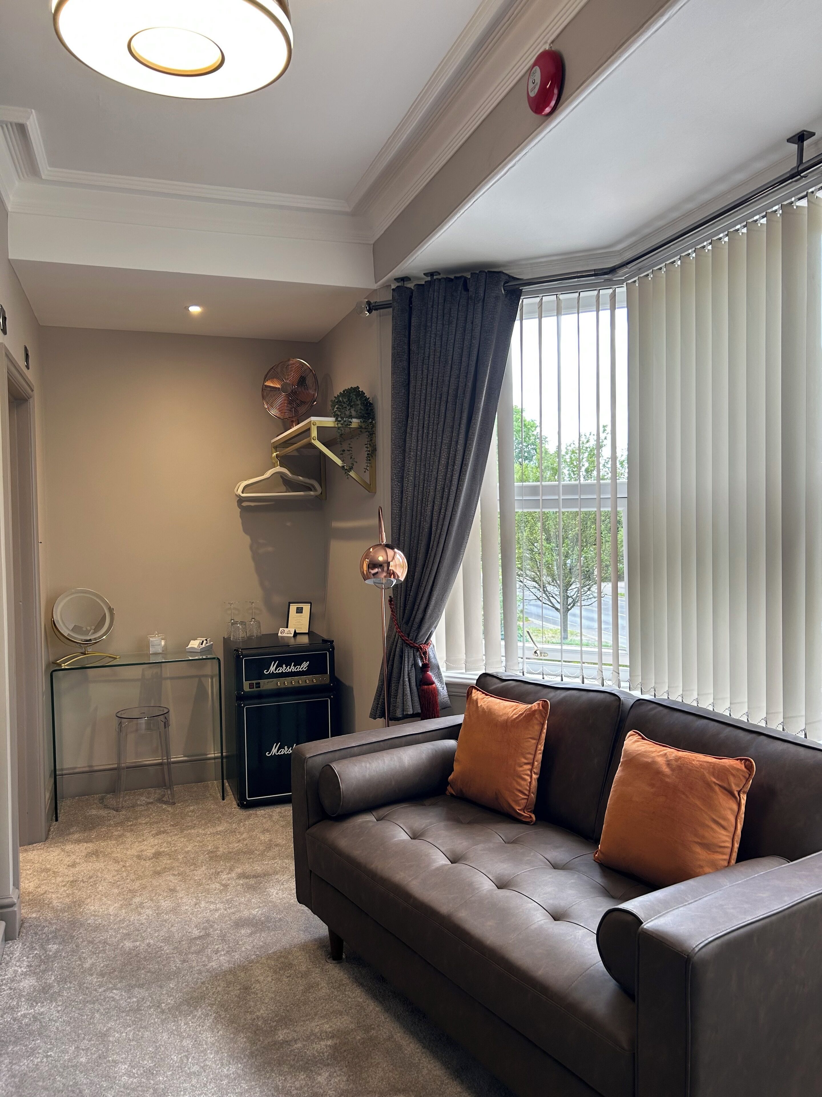 A modern living room with a leather sofa, a glass table, a Marshall amplifier, and a large window with blinds.