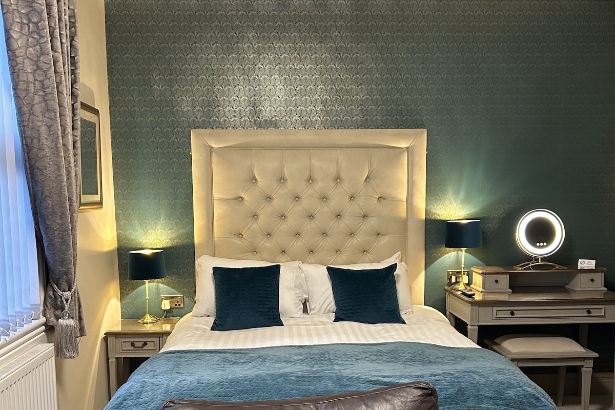 Un dormitorio ordenado con cabecera capitonada, cama con ropa de cama azul y blanca, sillón de cuero y mesita de noche con lámpara y espejo.