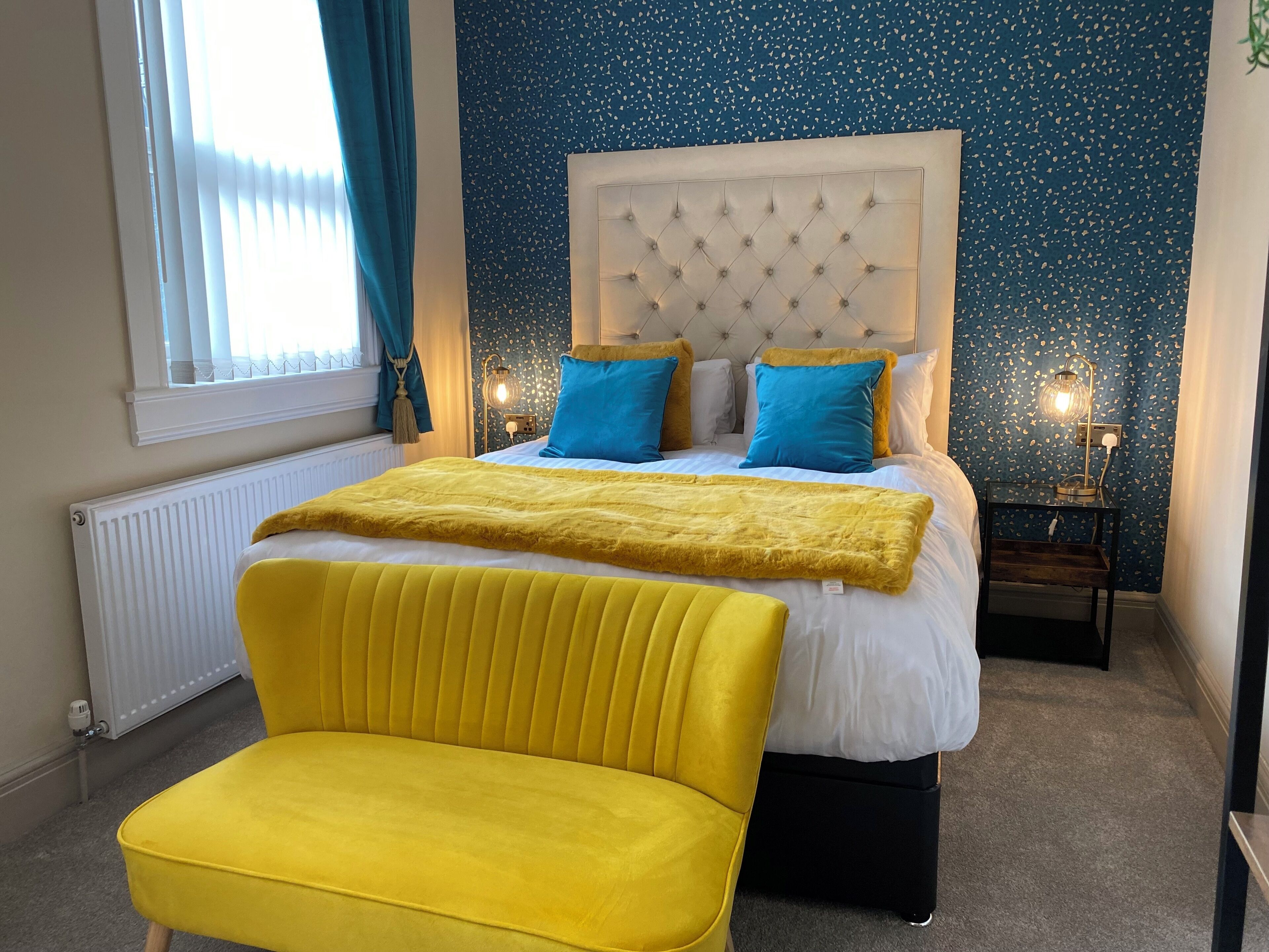 A bedroom with a tufted headboard, a yellow chair, a bed with blue and white bedding, and a nightstand with a lamp.