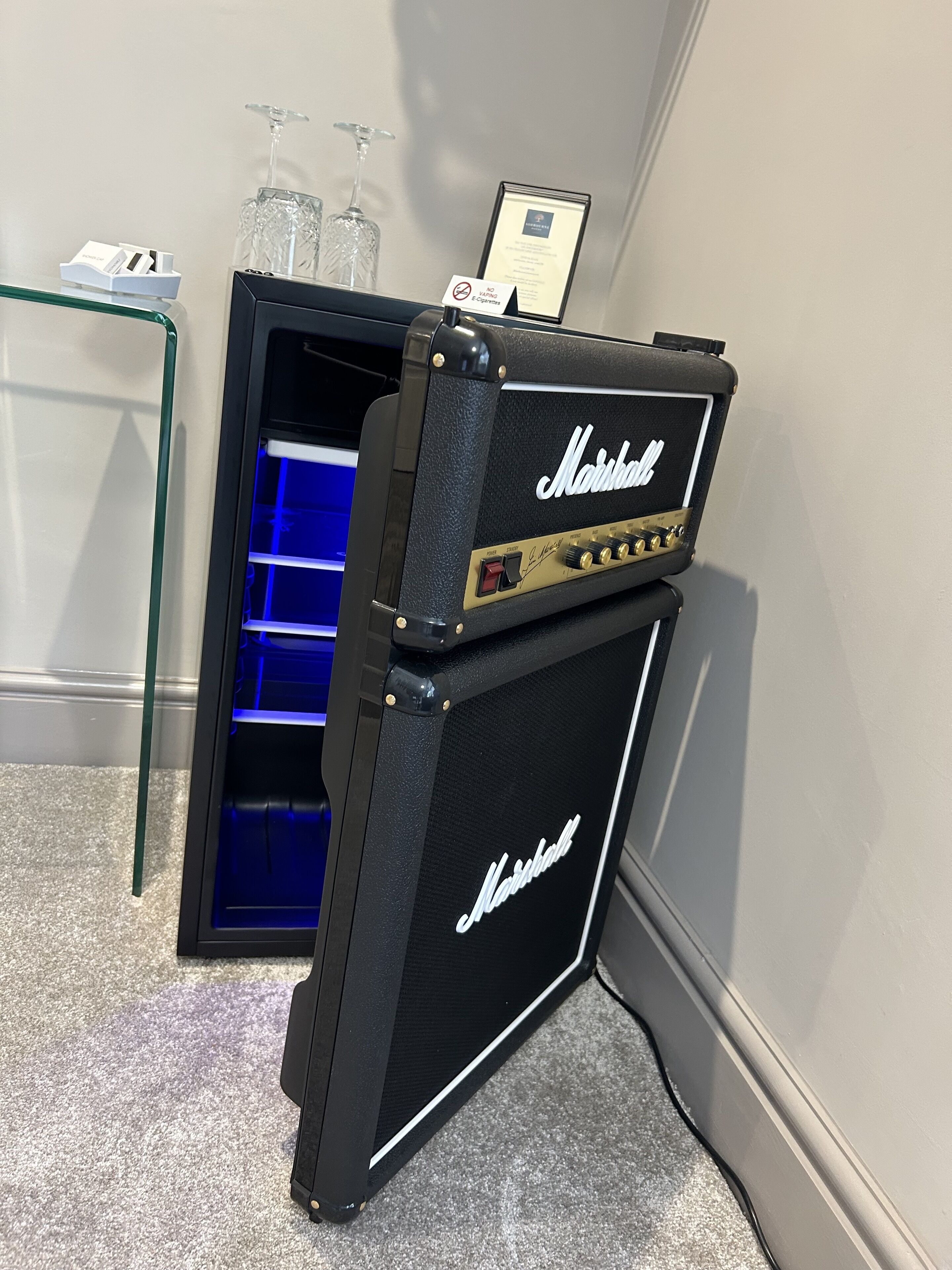 Un amplificador Marshall con un compartimento iluminado en azul, colocado en una esquina con una mesa de vidrio y un certificado enmarcado en la pared.