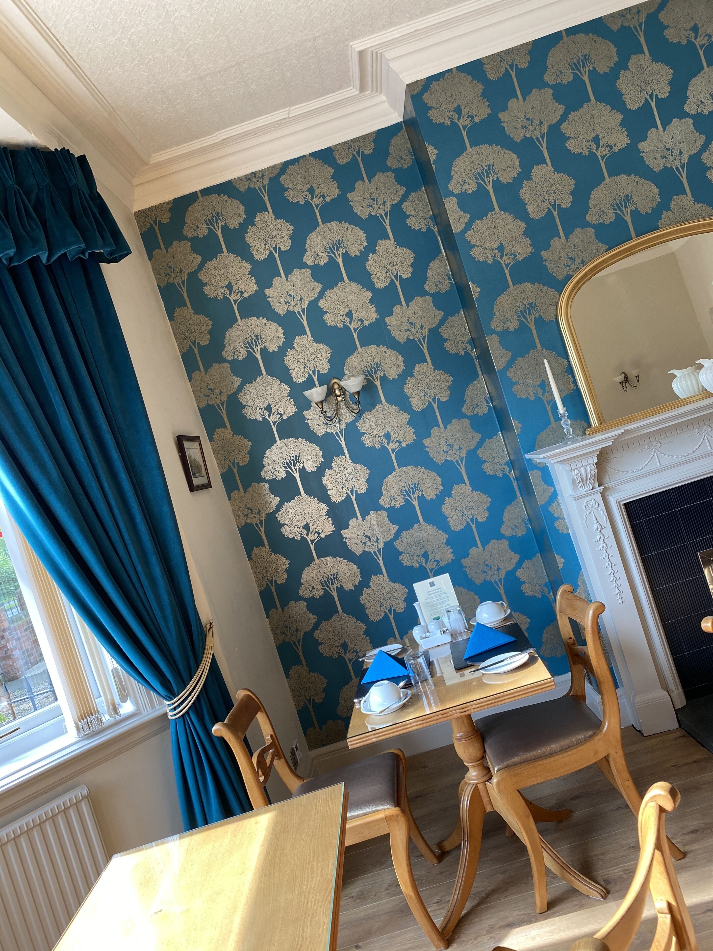 A dining area with a table set for two, a blue patterned wallpaper, a fireplace, and a window with curtains.