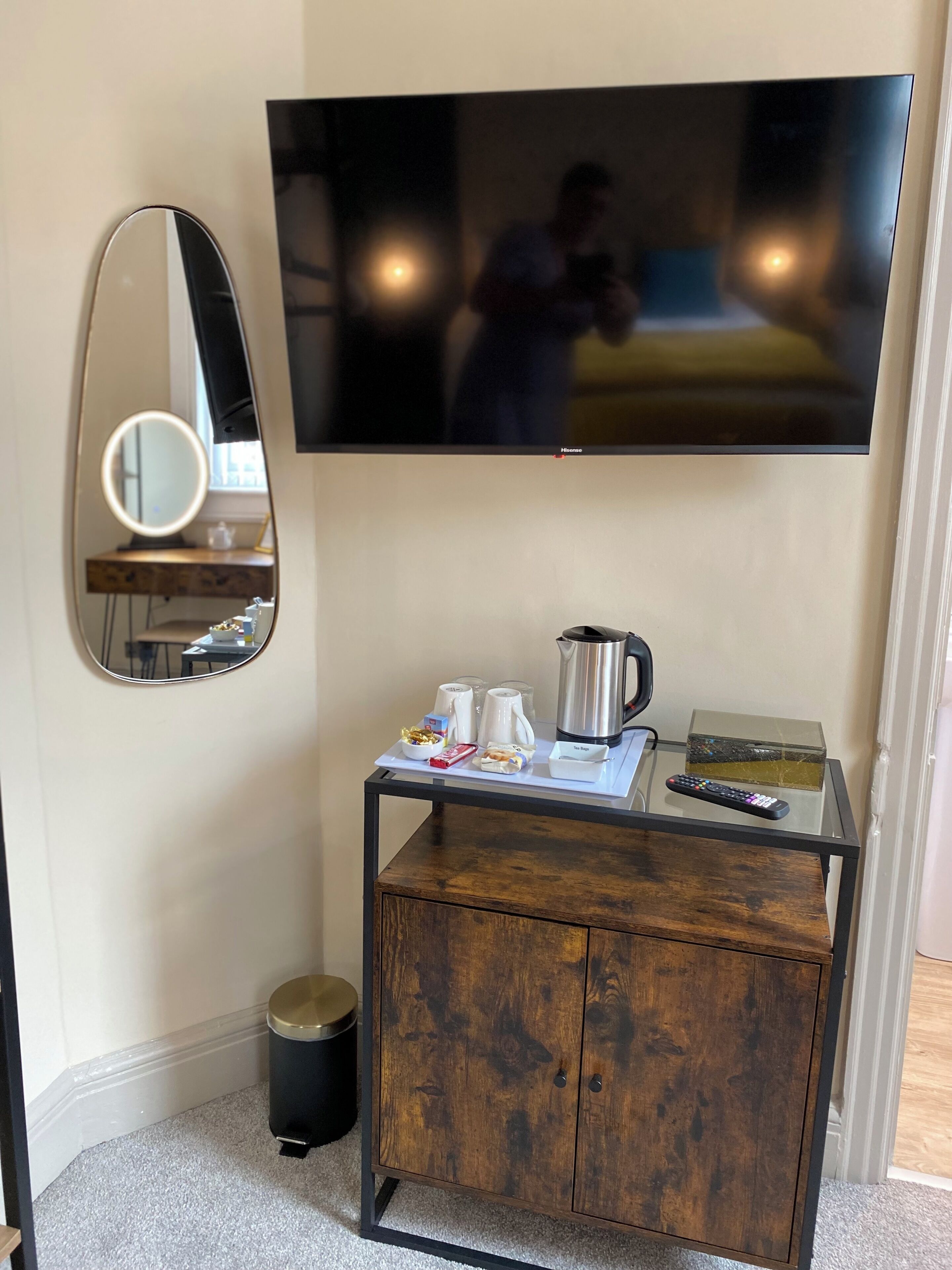 A room with a TV mounted on the wall, a wooden cabinet with a glass top, a mirror, and a trash bin.