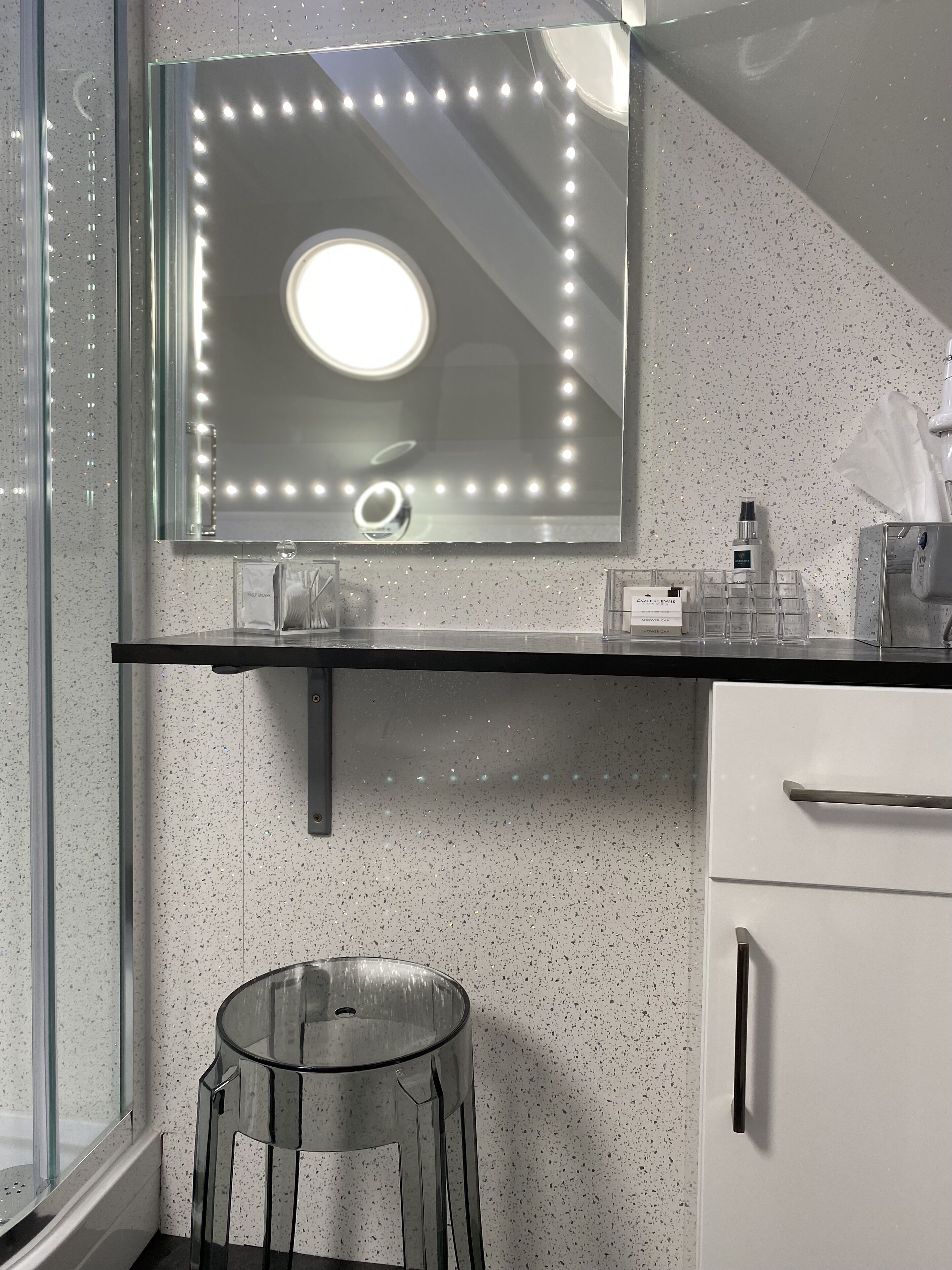 A bathroom with a vanity, a mirror with lights, a stool, and a hand dryer.