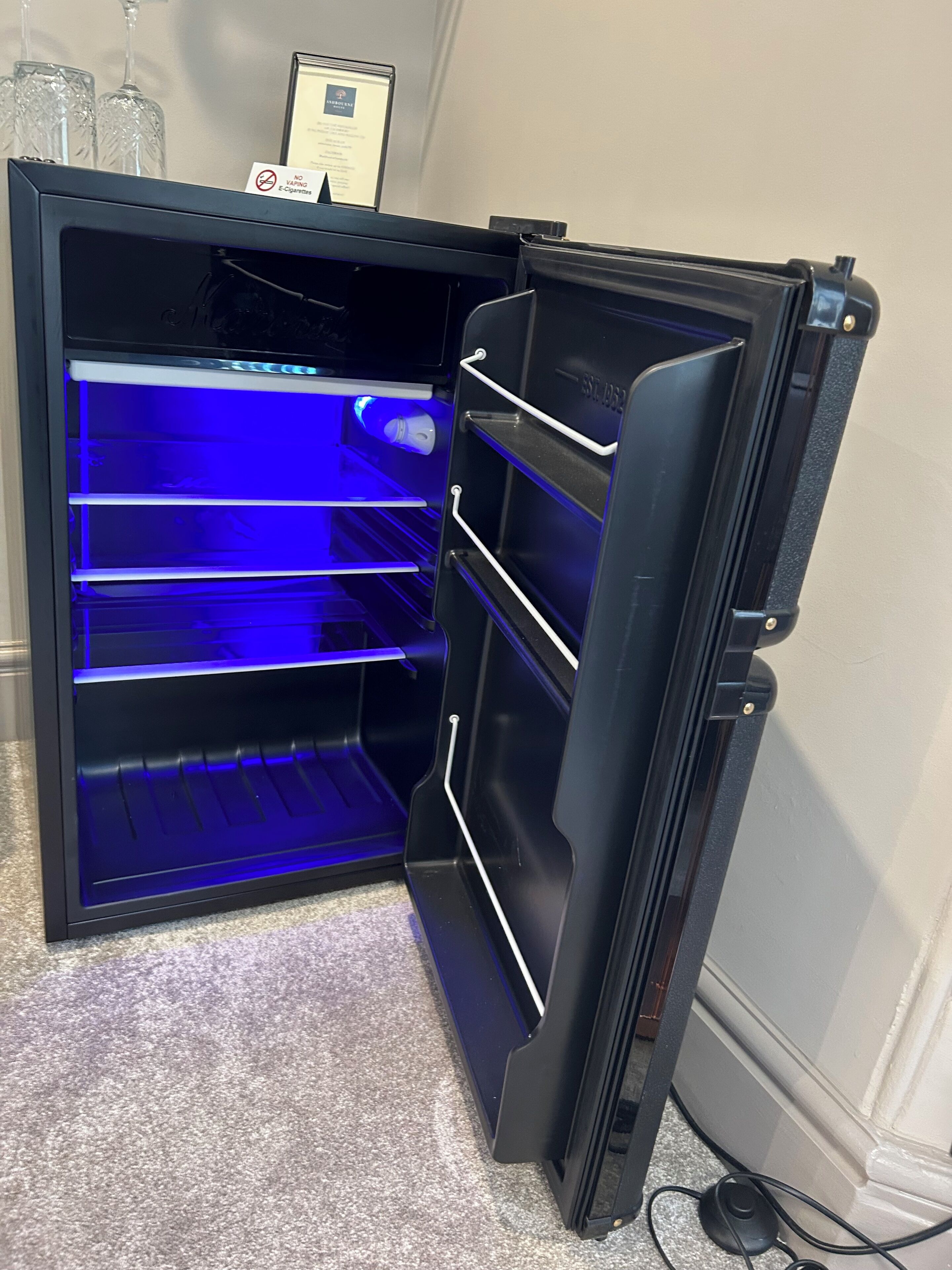 An open refrigerator with a blue light inside, a 'No Smoking' sign, and a framed certificate on top.