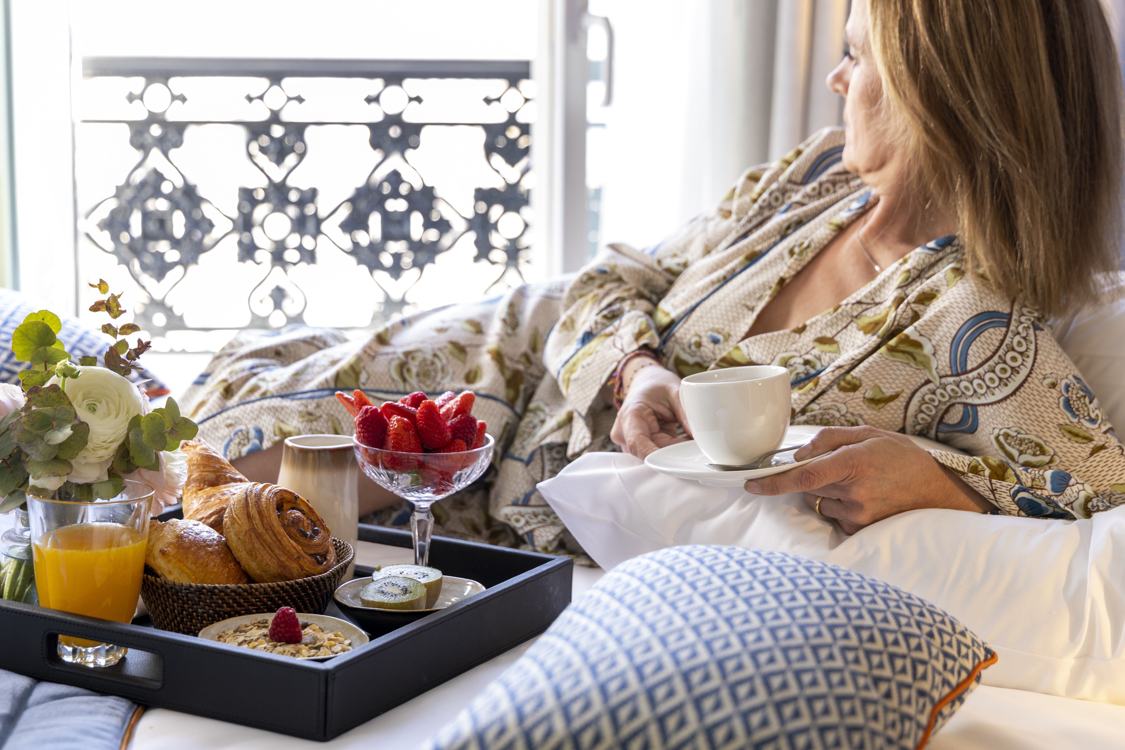 Eine Person in einem gemusterten Bademantel sitzt auf einem Bett mit einem Tablett mit Frühstücksleckereien, darunter Croissants, Erdbeeren und ein Glas Orangensaft.