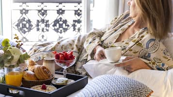 Eine Person in einem gemusterten Bademantel sitzt auf einem Bett mit einem Tablett mit Frühstücksleckereien, darunter Croissants, Erdbeeren und ein Glas Orangensaft.