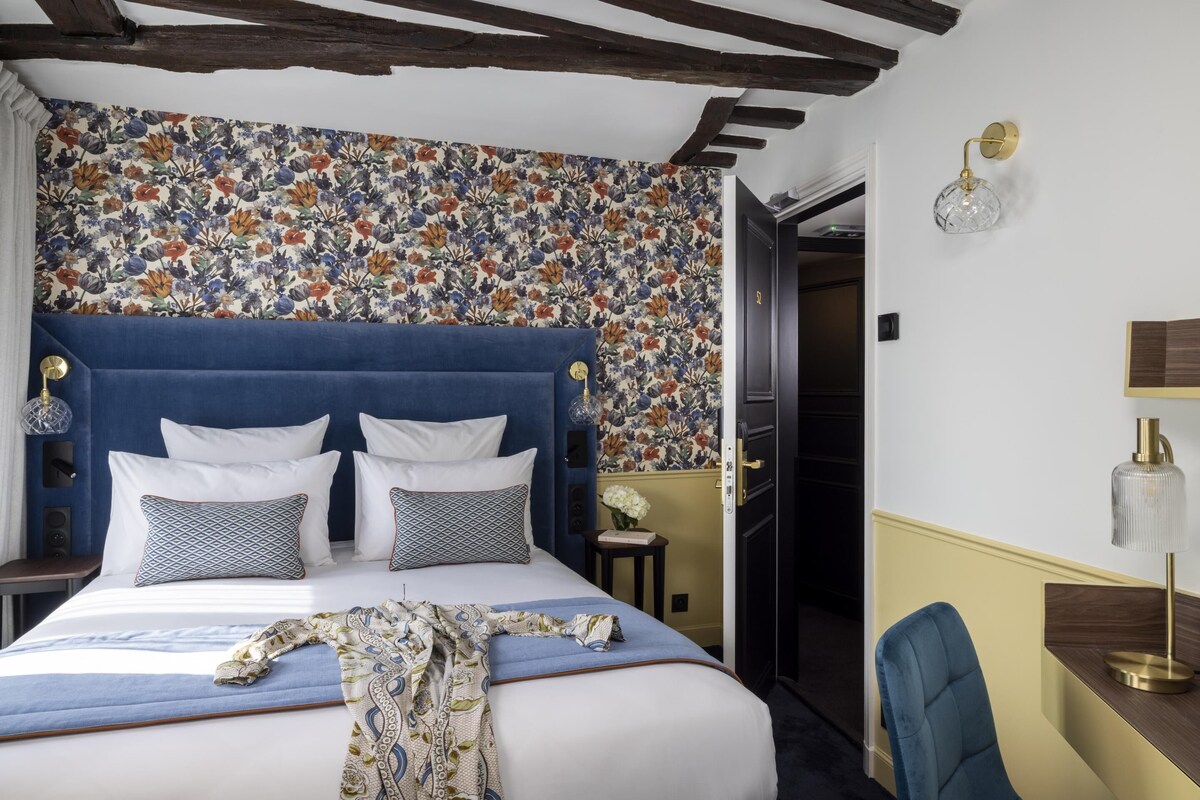 Ein Hotelzimmer mit einem großen Bett, floralem Wanddesign, einem blauen, gepolsterten Kopfteil und einem Schreibtisch mit Lampe.