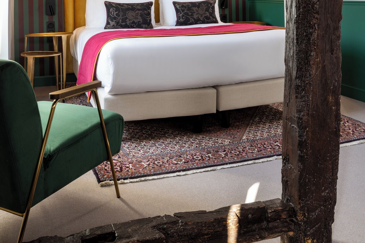 Ein Schlafzimmer mit schrägem Dach, einem Bett mit weißer Bettwäsche, einem grünen Sessel und einem gemusterten Teppich.
