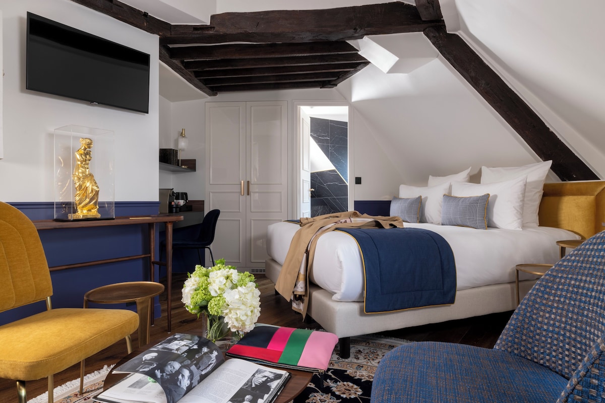 Ein Schlafzimmer mit Bett, Nachttisch und Schreibtisch mit Stuhl. Ein Fernseher ist an der Wand montiert, und ein Fenster sorgt für natürliches Licht.