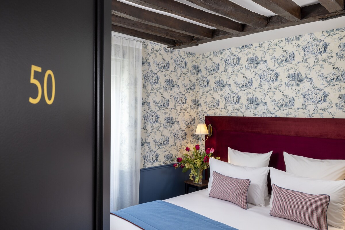 Ein Hotelzimmer mit Türschild Nummer 50, floralem Wanddesign, einem roten Kopfteil, weißer Bettwäsche und einem Nachttisch mit Lampe und Blumen.