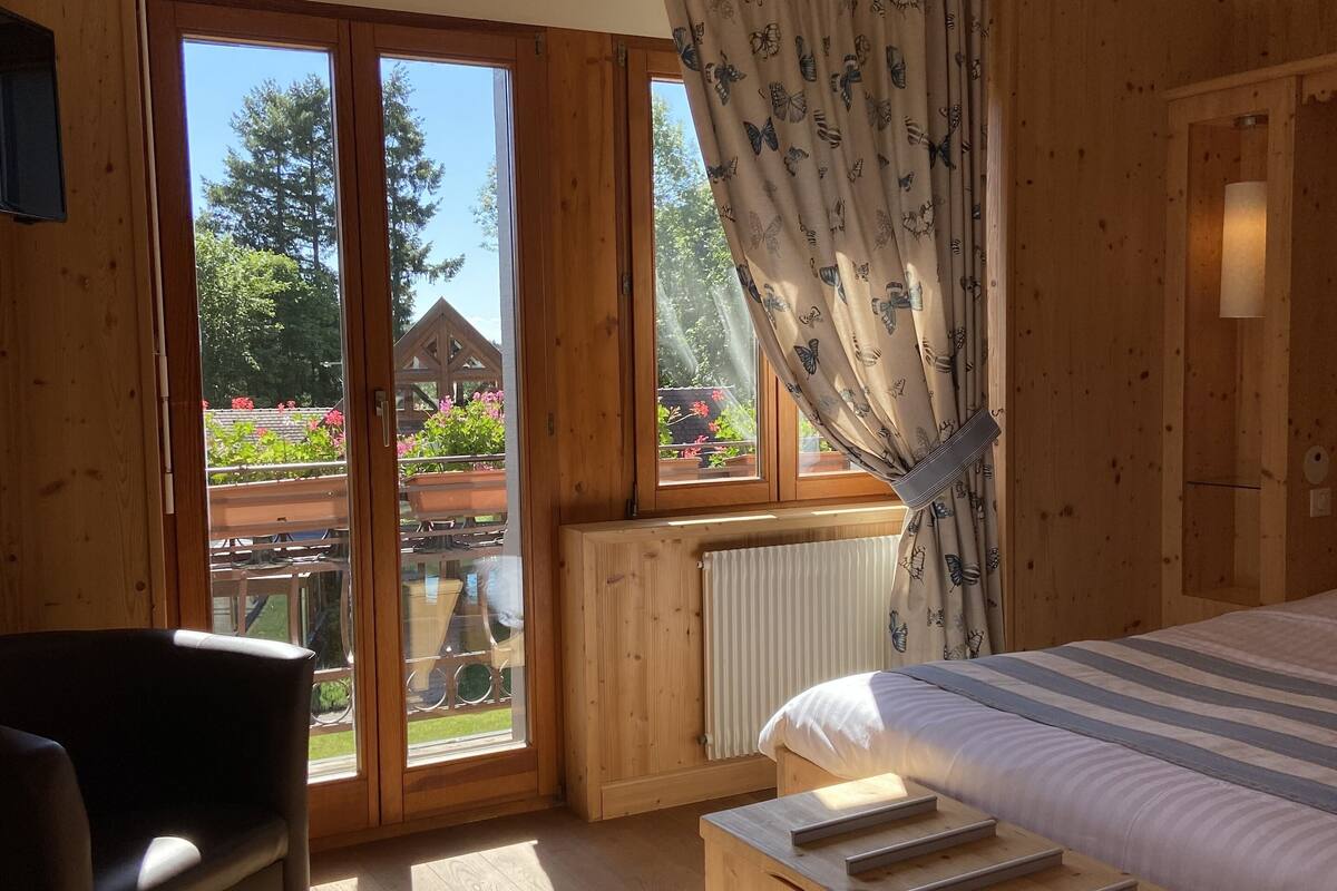 A wooden cabin room with a bed, a chair, and a view of a garden through large windows.