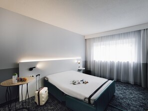 A hotel room with a bed, a small table, a suitcase, a lamp, and a window with curtains.