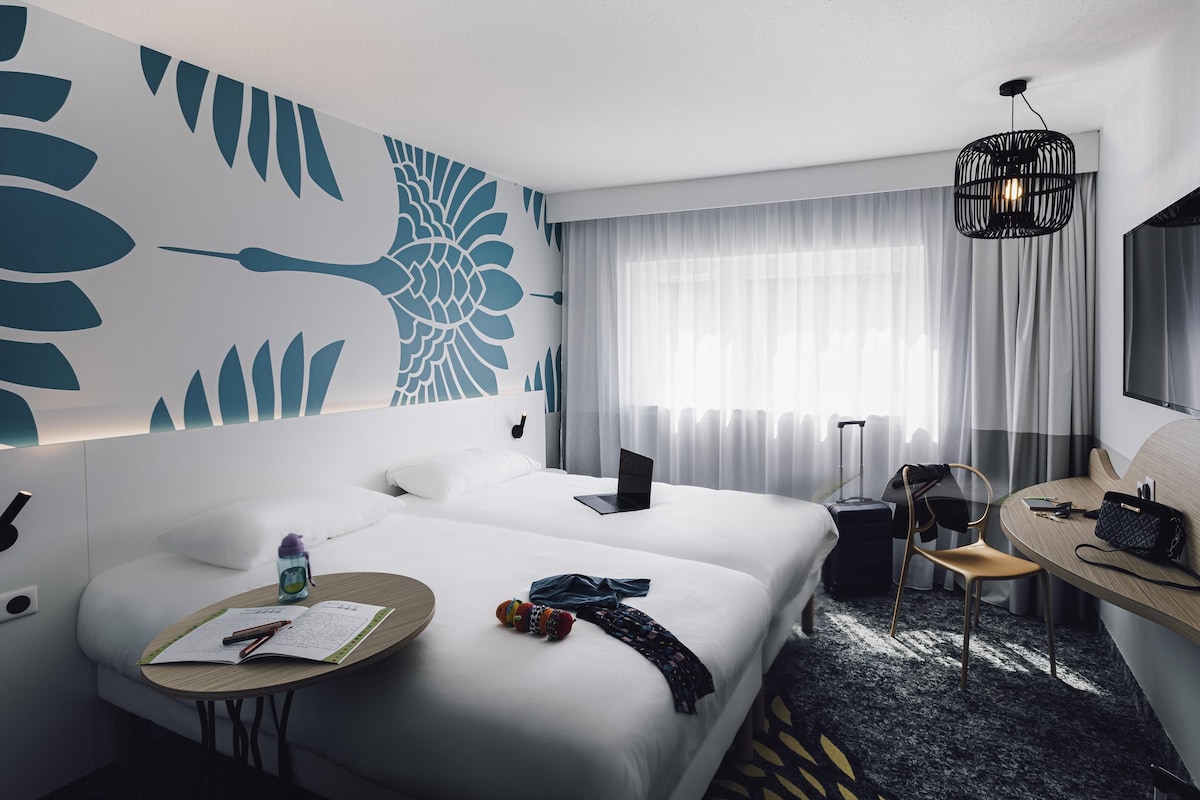 Habitación de hotel con cama, mesita de noche, silla, escritorio y un gran ventanal con cortinas.