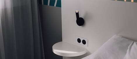A modern hotel room with a minimalist design, featuring a white bedside table, a small white nightstand, and a wall-mounted light fixture.