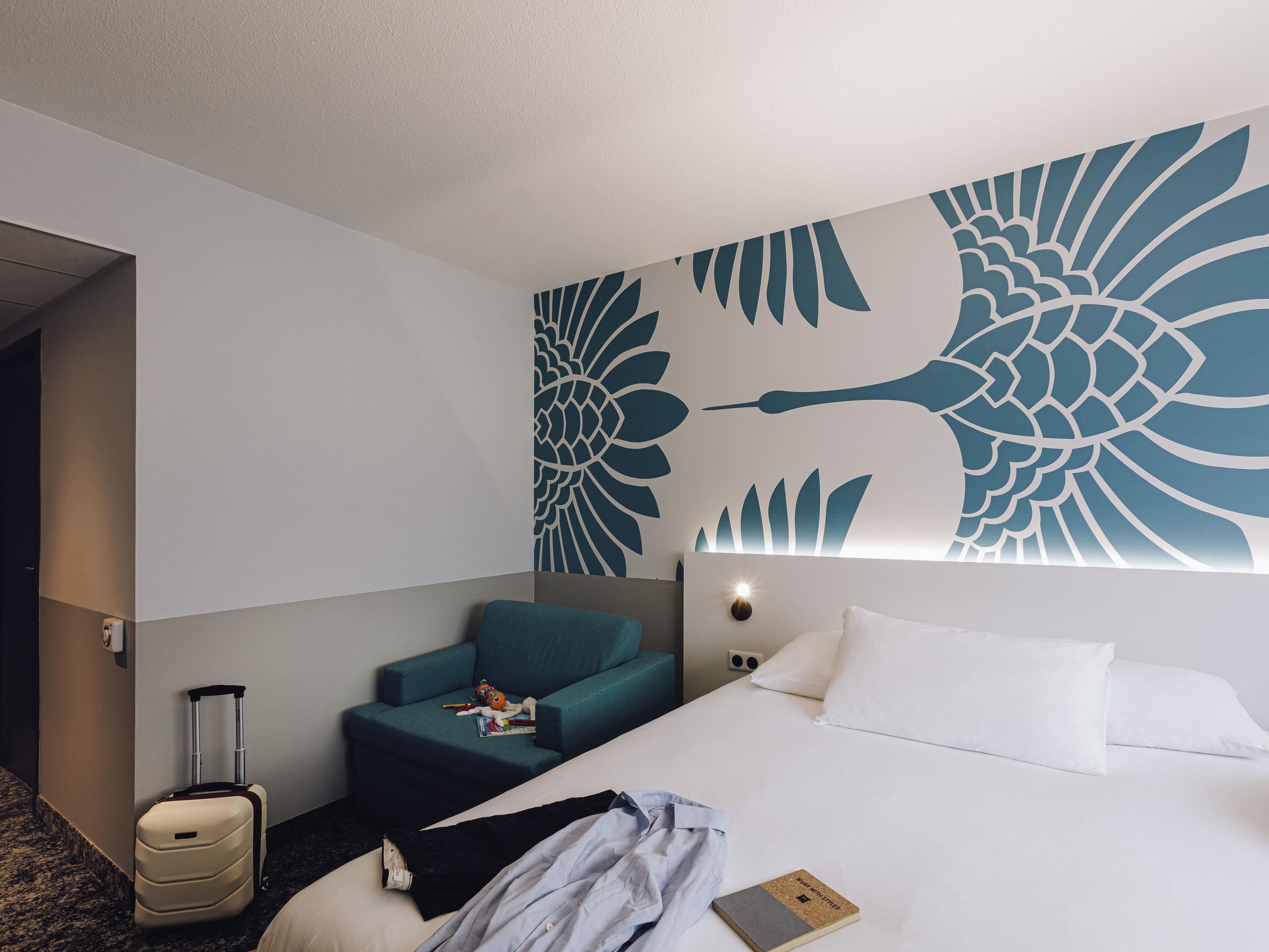 A hotel room with a bed, a chair, a suitcase, and a wall with a large floral design.