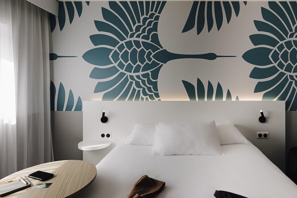 A hotel room with a bed, bedside tables, a wall with a large floral pattern, a window with curtains, and a small round table with a phone and a wallet.
