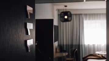 Una habitación de hotel moderna con una puerta de madera oscura, un escritorio con silla, una cama y un amplio ventanal con cortinas.