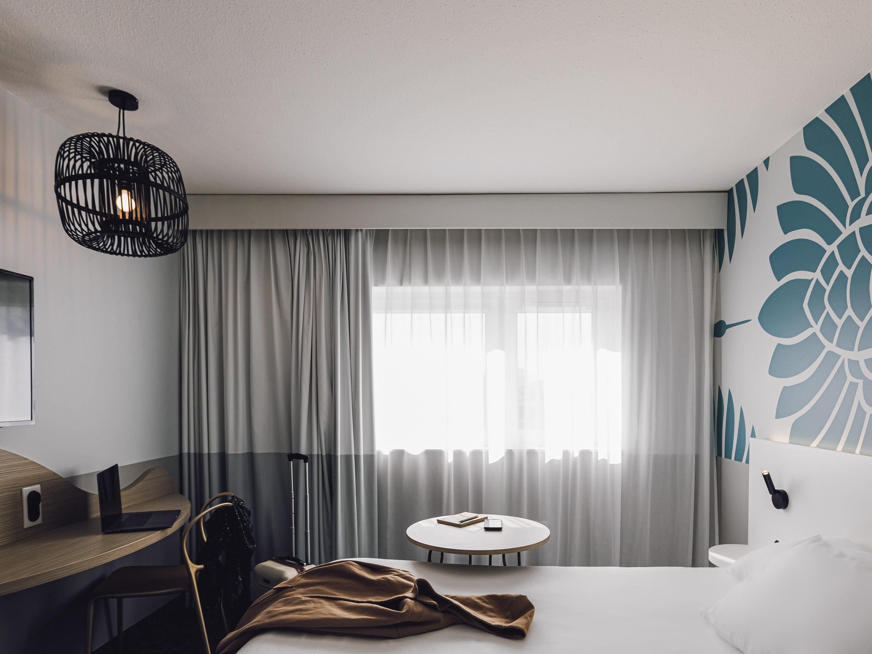 Habitación de hotel con cama, escritorio con una computadora portátil, silla, mesa redonda y un ventanal con cortinas.