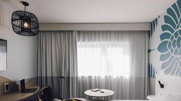 Habitación de hotel con cama, escritorio con una computadora portátil, silla, mesa redonda y un ventanal con cortinas.