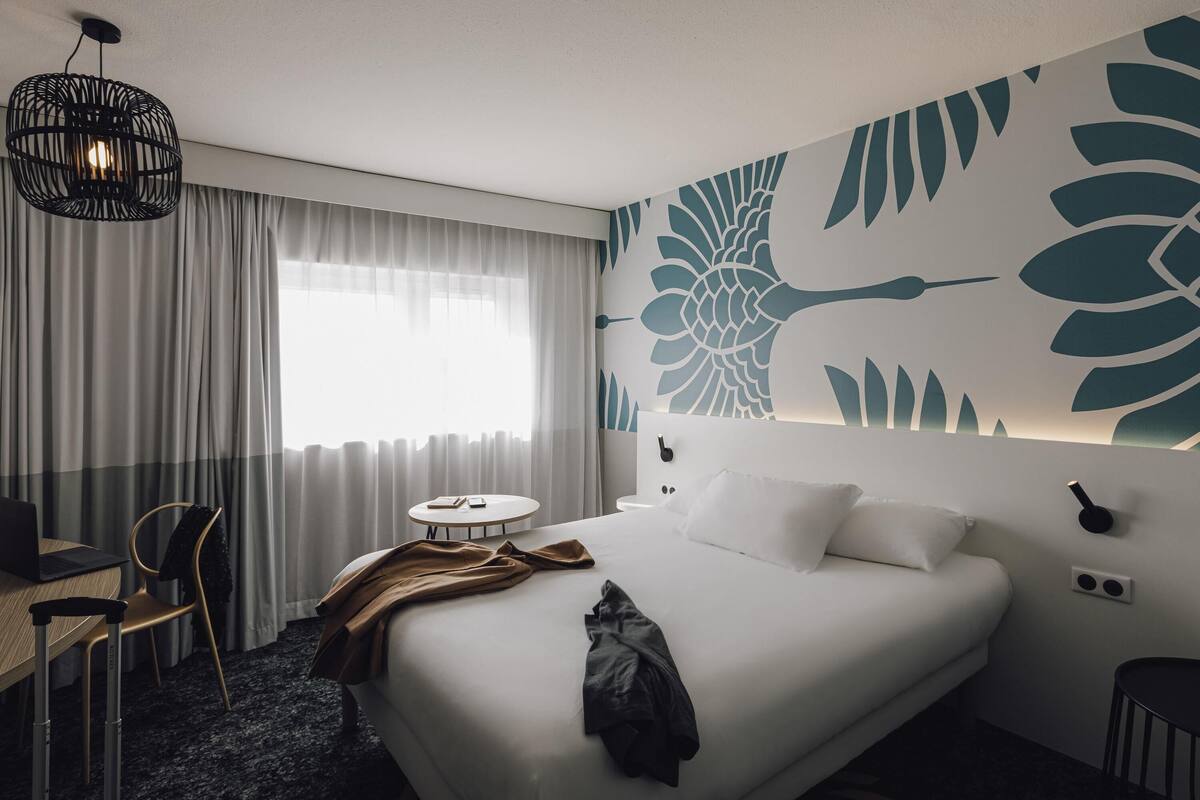 Une chambre d’hôtel comprenant un lit, un bureau avec un ordinateur portable, une chaise, une table ronde et un mur orné d’un motif floral.