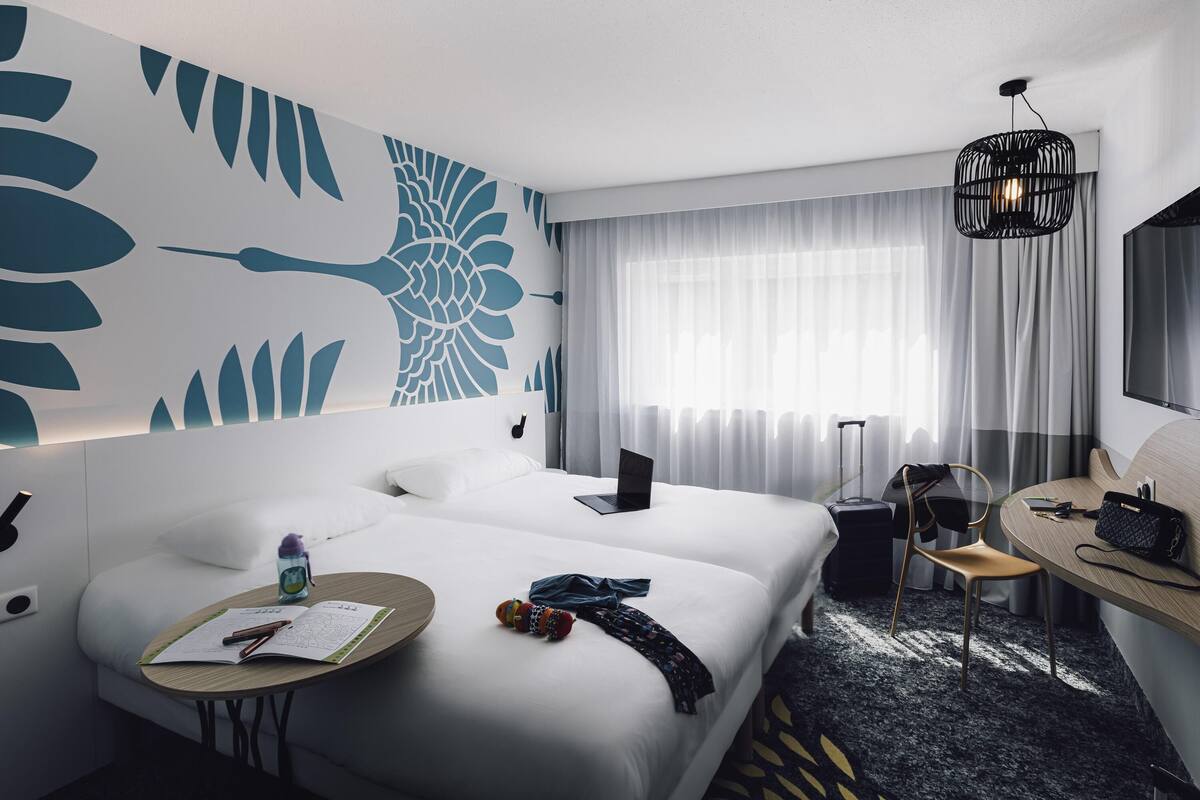 Une chambre d’hôtel comprenant un lit, une table de chevet, une chaise, un bureau et une grande fenêtre avec des rideaux.