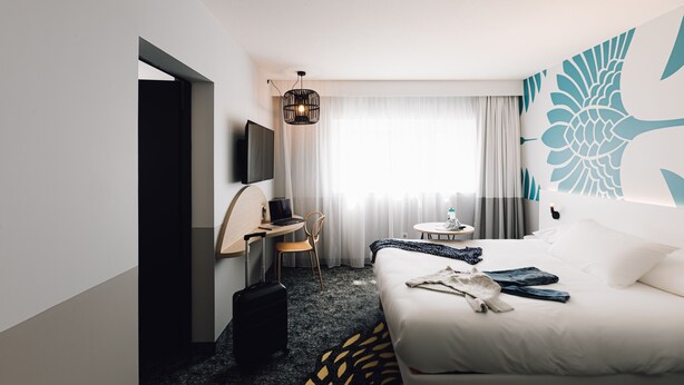 A hotel room with a bed, a desk, a chair, a suitcase, a TV, and a window with curtains. - ibis Styles Nancy Laxou (Laxou)